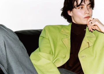 Model wearing a bright green blazer and dark turtleneck leaning against a dark surface against a white wall.