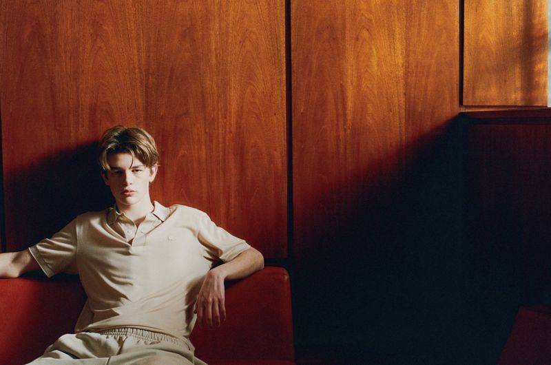 Young adult male in polo shirt and sweatpants seated on red couch against wood panel wall in indoor setting with natural light.