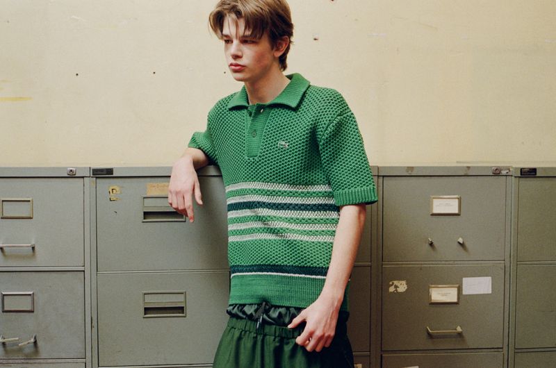 Young man in green Lacoste knit polo shirt stands in front of grey metal file cabinet for a fashion editorial shot in a studio.