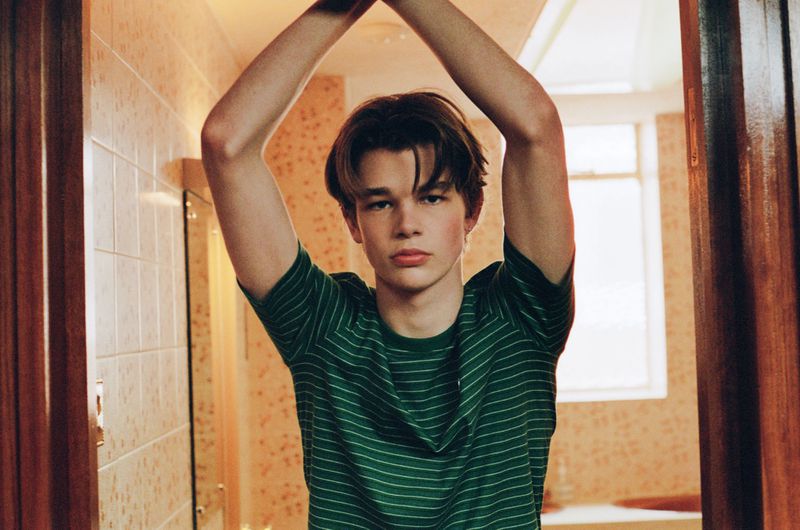Indoor portrait of a young adult male with brown hair, wearing a green striped shirt, posing with arms raised in a tiled bathroom.