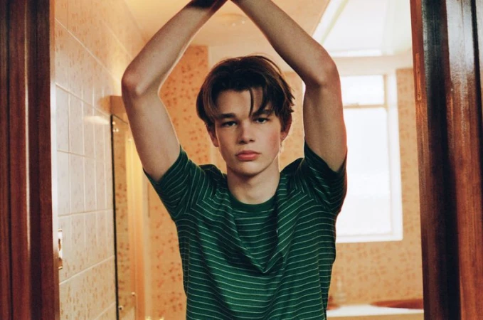 Young man in a green striped t-shirt standing inside a tiled interior doorway with arms raised