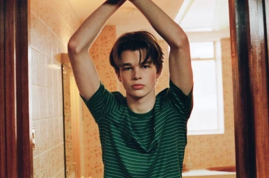 Young man in a green striped t-shirt standing inside a tiled interior doorway with arms raised