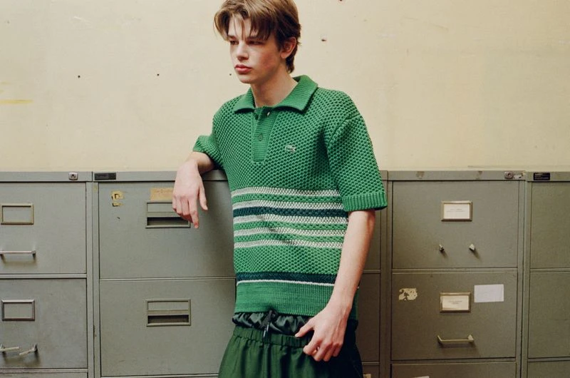 Young male model leaning against grey metal filing cabinets wearing a textured green polo shirt and dark trousers.