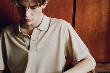 Young man wearing a cream polo shirt seated against a wood-paneled wall for the LN-CC x Lacoste project