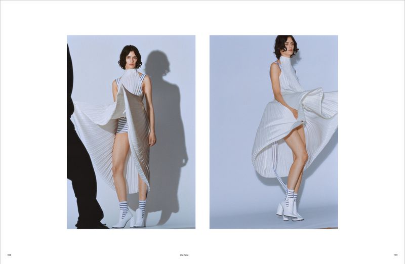 Fashion shot of a woman in a white pleated dress and white boots, posing in front of a light blue studio backdrop with soft lighting.