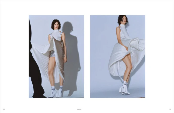 Fashion shot of a woman in a white pleated dress and white boots, posing in front of a light blue studio backdrop with soft lighting.