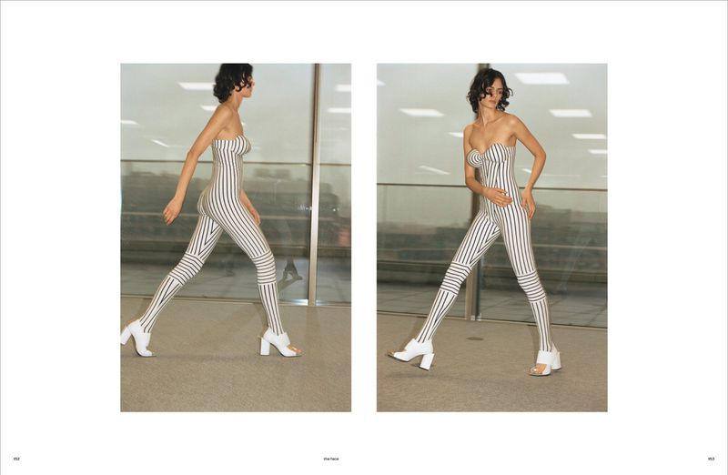 A slender woman models a full-body striped strapless garment and white block-heeled shoes in a minimal indoor setting with natural light.