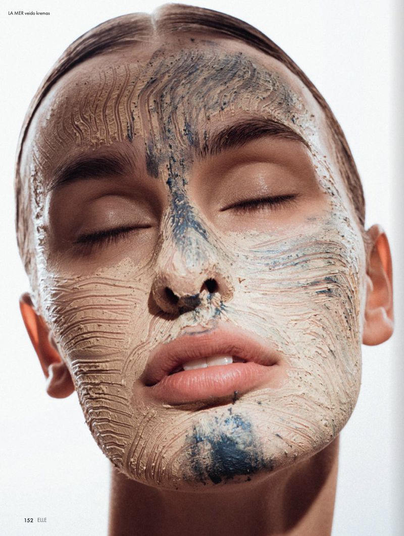 Close-up portrait of a woman with her eyes closed, featuring a facial mask with contrasting beige and blue clay textures on her skin.