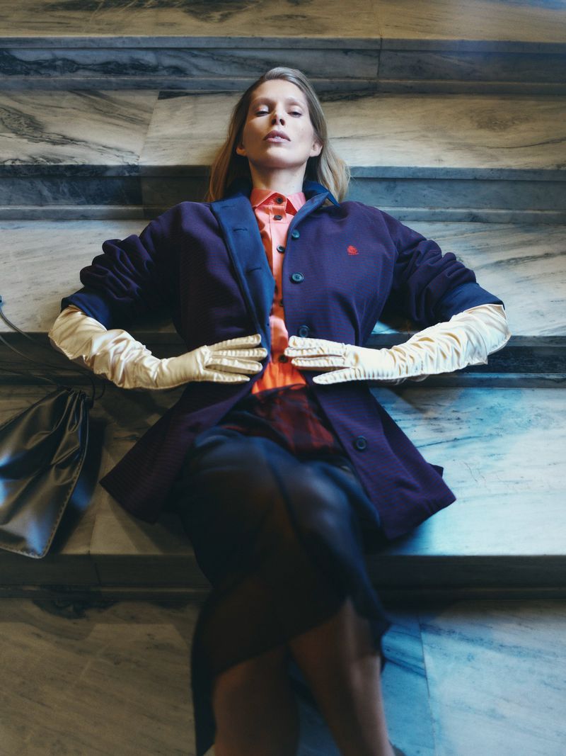 A blonde woman in a coat and gloves lies on marble stairs with a black purse. Shot from overhead. Fashion, beauty, style.