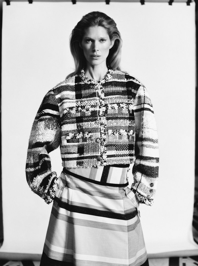 Monochrome fashion shot of an adult female model with blonde hair wearing a patterned jacket and striped skirt posing in a studio.