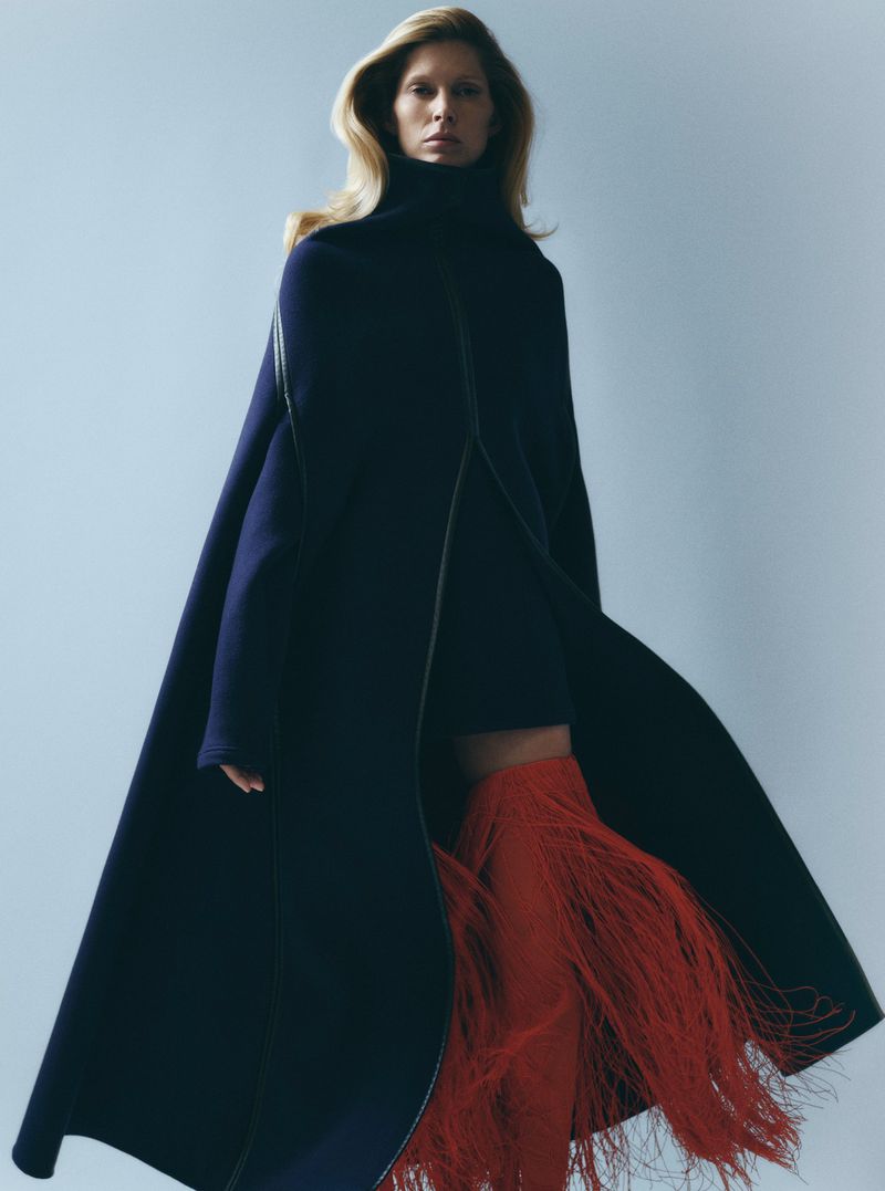 Fashion shot of an adult woman in a studio wearing a dark coat, short dress, and red fringe boots against a pale blue background.