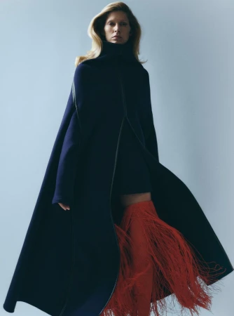 Fashion shot of an adult woman in a studio wearing a dark coat, short dress, and red fringe boots against a pale blue background.