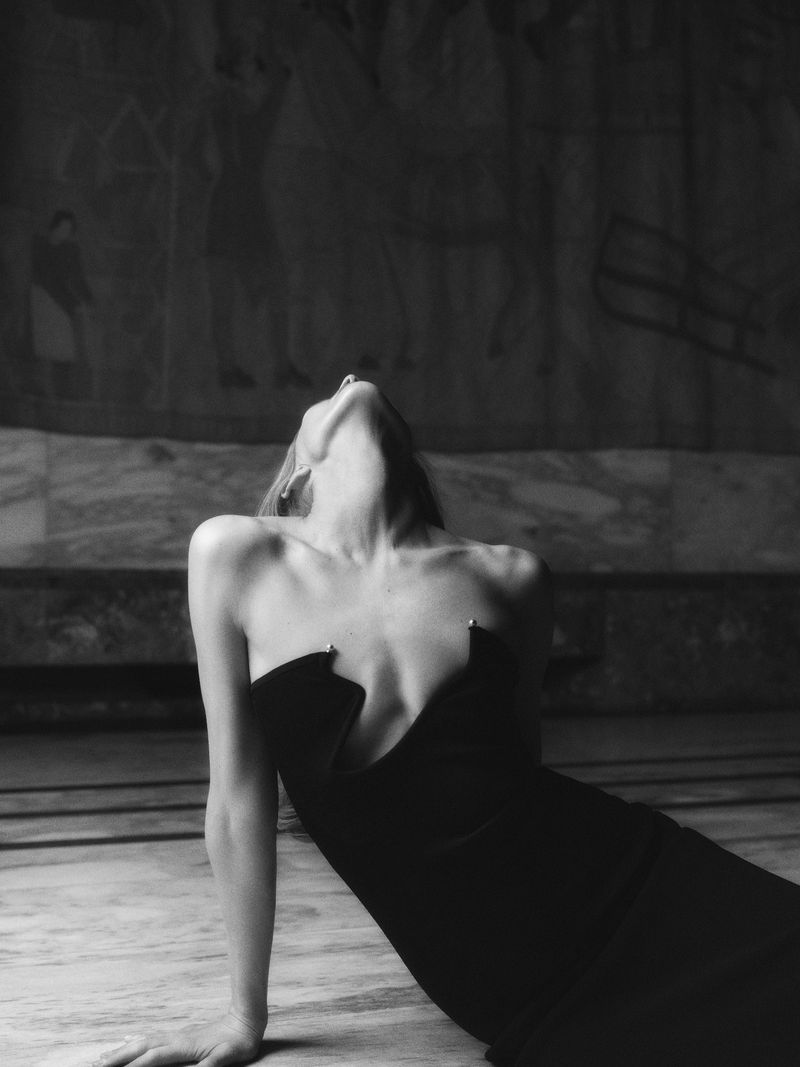 Black and white fashion portrait of a woman in a black strapless dress, reclining on a marble floor in front of a textured mural.