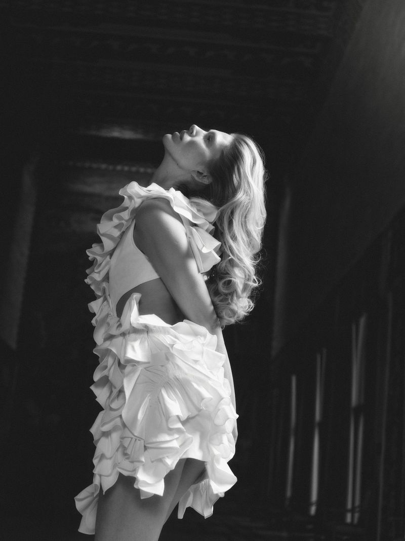 Black and white shot of a fair-skinned woman in a ruffled white dress, looking upwards in a dark interior setting, creating a high-fashion image.