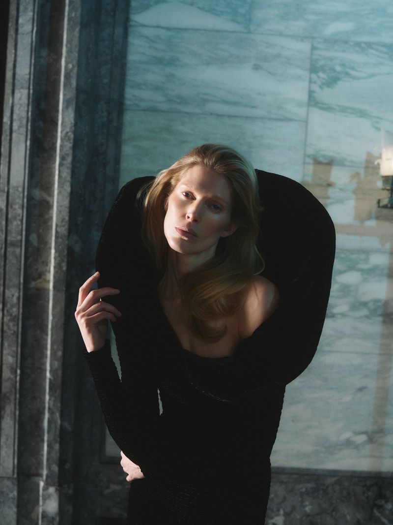 A woman in a black dress stands in front of a marble wall, with a dramatic black collar framing her face in an editorial fashion shot.