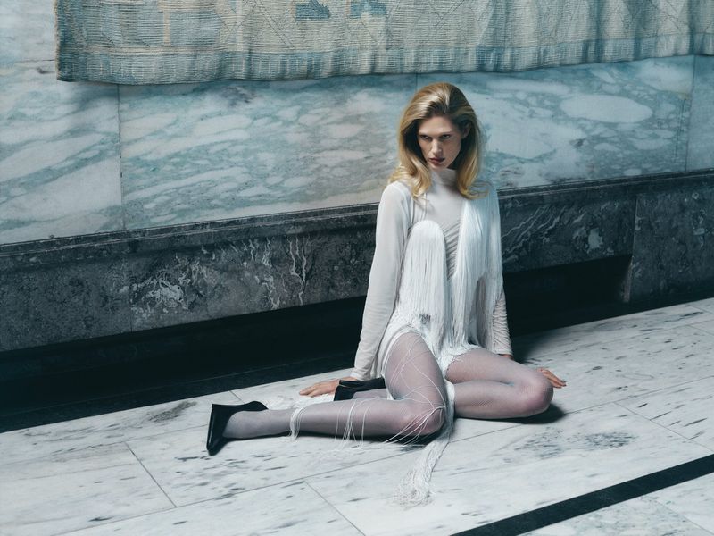 Blonde woman in sheer white dress with fringe detail poses on marble floor against a marble wall, wearing black heels.
