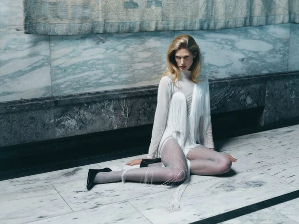 Blonde woman in sheer white dress with fringe detail poses on marble floor against a marble wall, wearing black heels.