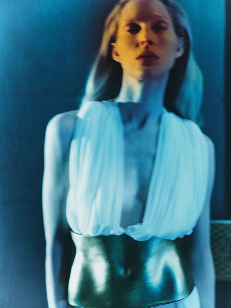 Portrait of a blonde woman in a flowing white dress with a metallic gold belt, set against a backdrop of cool blue tones and soft lighting.
