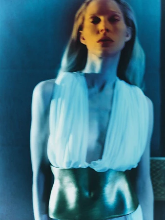 Portrait of a blonde woman in a flowing white dress with a metallic gold belt, set against a backdrop of cool blue tones and soft lighting.