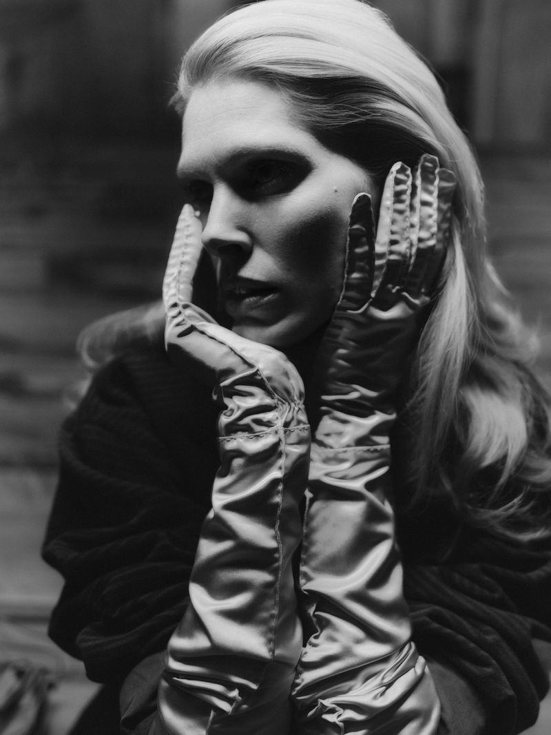 Black and white portrait of a blond woman wearing opera-length gloves and resting her hands on her face in a studio setting.