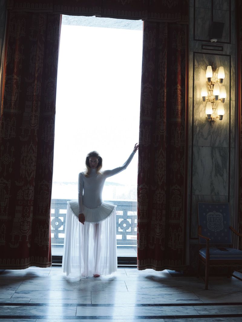 Elegant woman in white dress stands by window with drawn curtains, framed by marble wall and glowing wall sconces in a serene indoor scene.