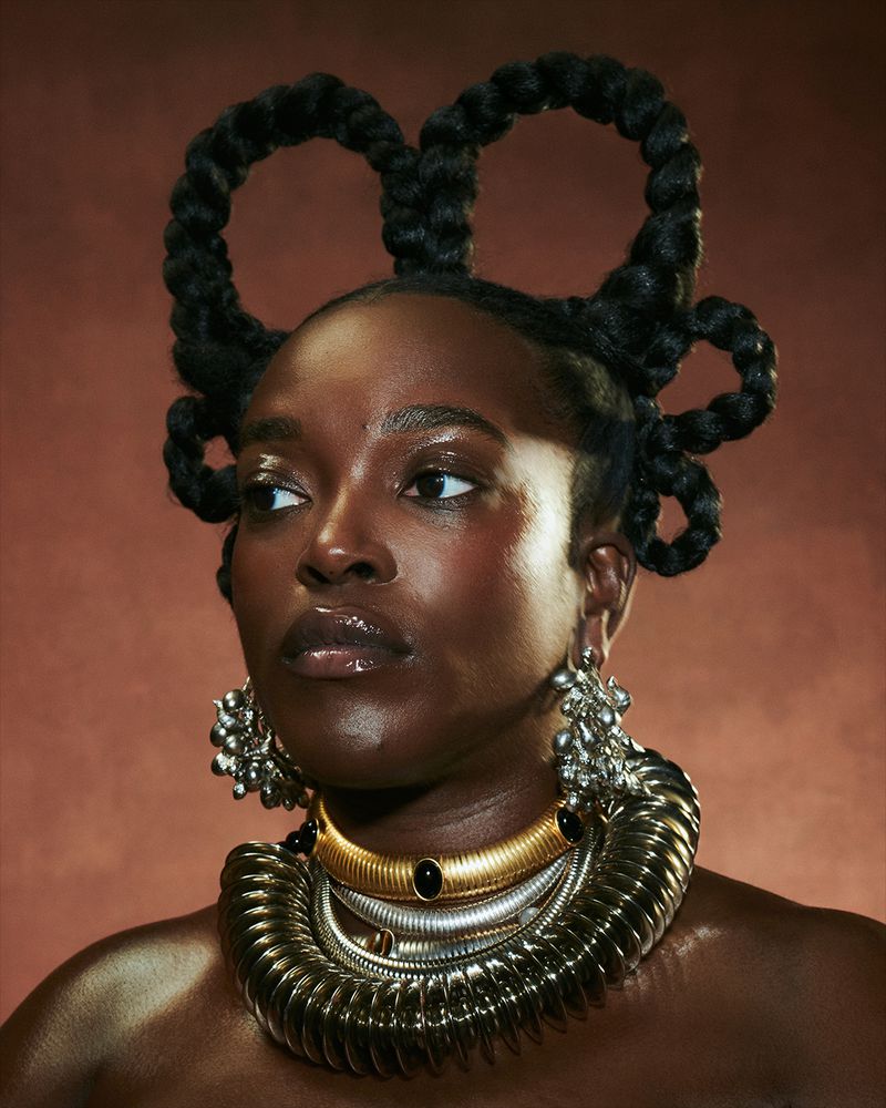 Portrait of a Black woman with dark skin, a unique hairstyle, and wearing multiple gold and silver necklaces and silver earrings on a brown backdrop.