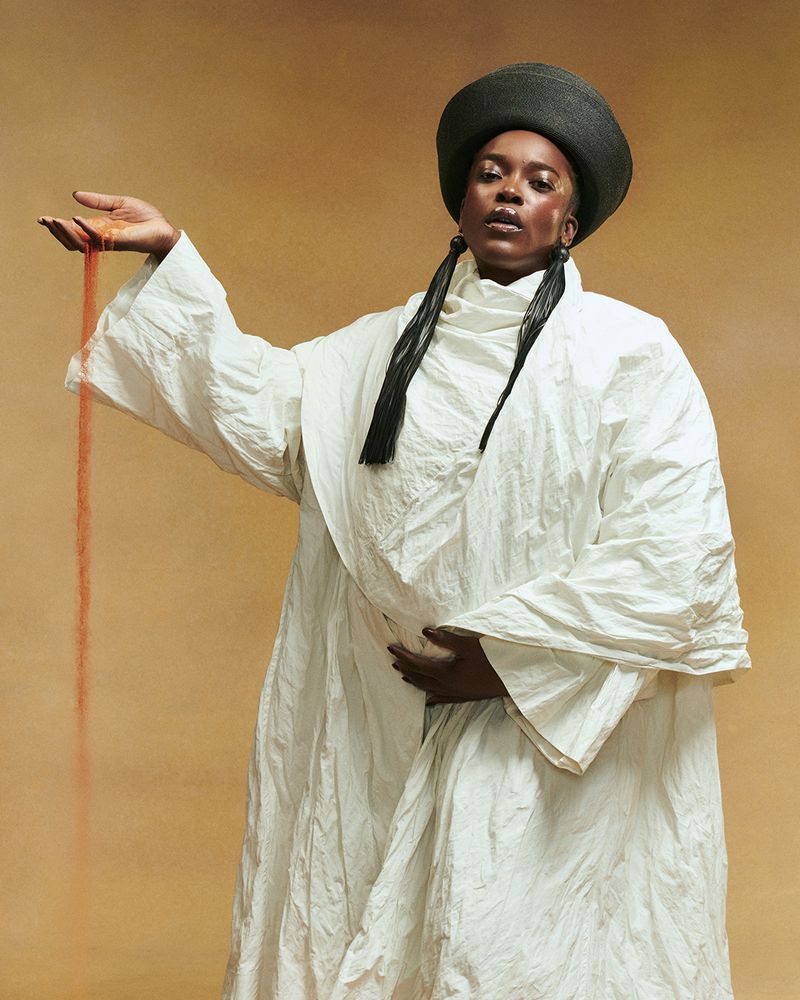 Woman in white robe poses with sand in hand against tan backdrop in studio. Long black earrings, simple wardrobe, portrait orientation.