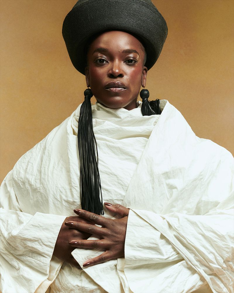 Headshot of an adult Black woman in a black hat and flowing white top against a tan background. The woman has long tassel earrings.