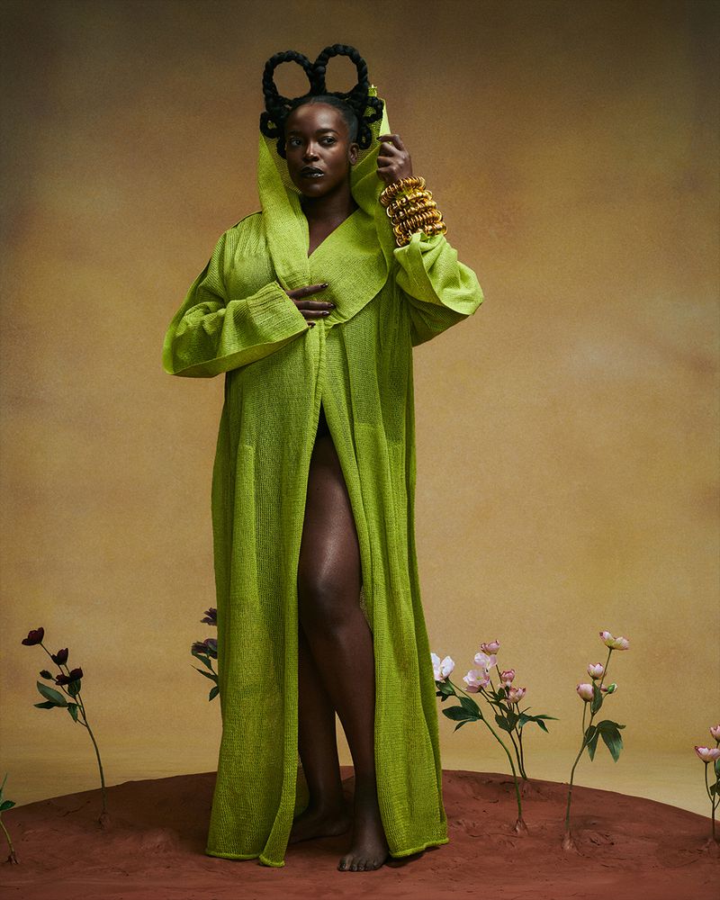 Full-length portrait of an adult Black woman wearing a hooded green gown and golden bracelets, standing on a clay-like surface with flowers.