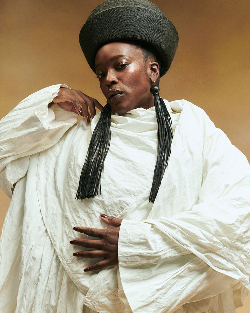 Headshot of a Black woman wearing a large textured hat, statement tassel earrings, and draped white clothing against a neutral backdrop.