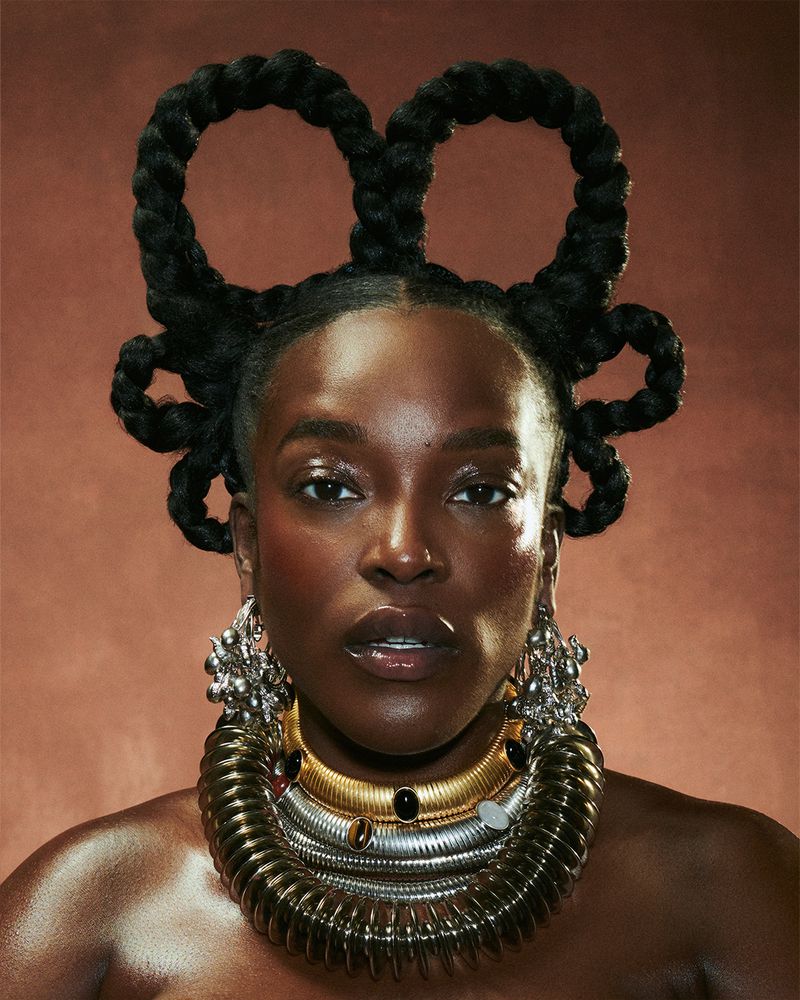 Close-up portrait of an adult woman with dark skin, braided hair loops, and elaborate silver and gold jewelry against a brown background.