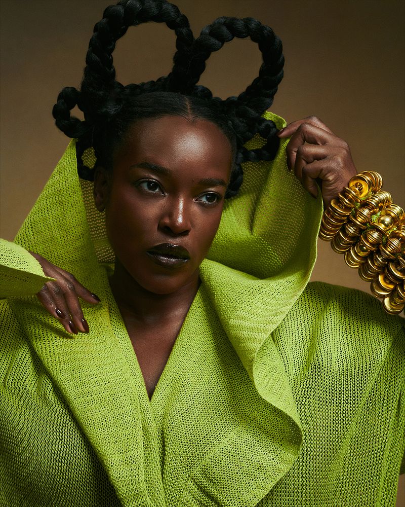 Adult female poses in a hooded chartreuse garment, adorned with a large gold bracelet, against a neutral backdrop in a studio shot.