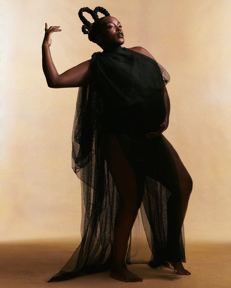 Pregnant Black woman in sheer black fabric poses in a studio with a beige background. She is standing barefoot with her hands on her belly.