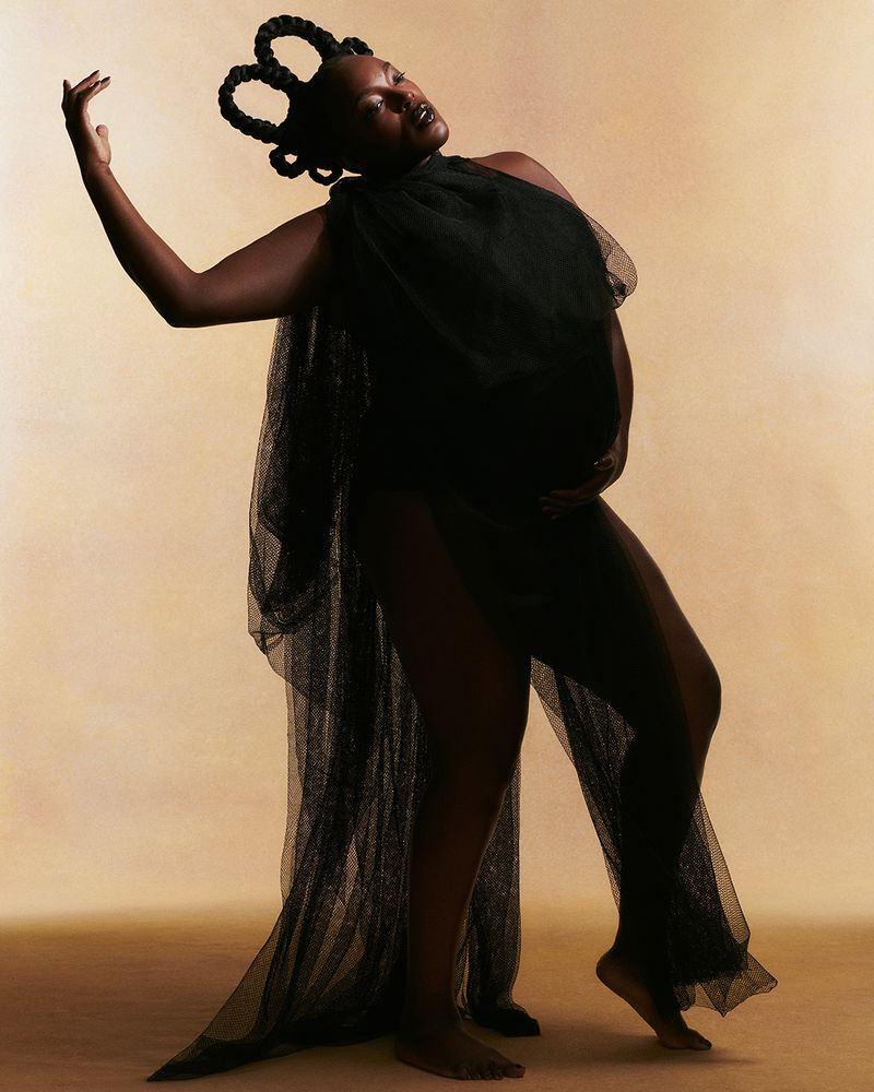 Pregnant Black woman with braided hair, wearing a black dress, posing against a tan backdrop in a studio setting.