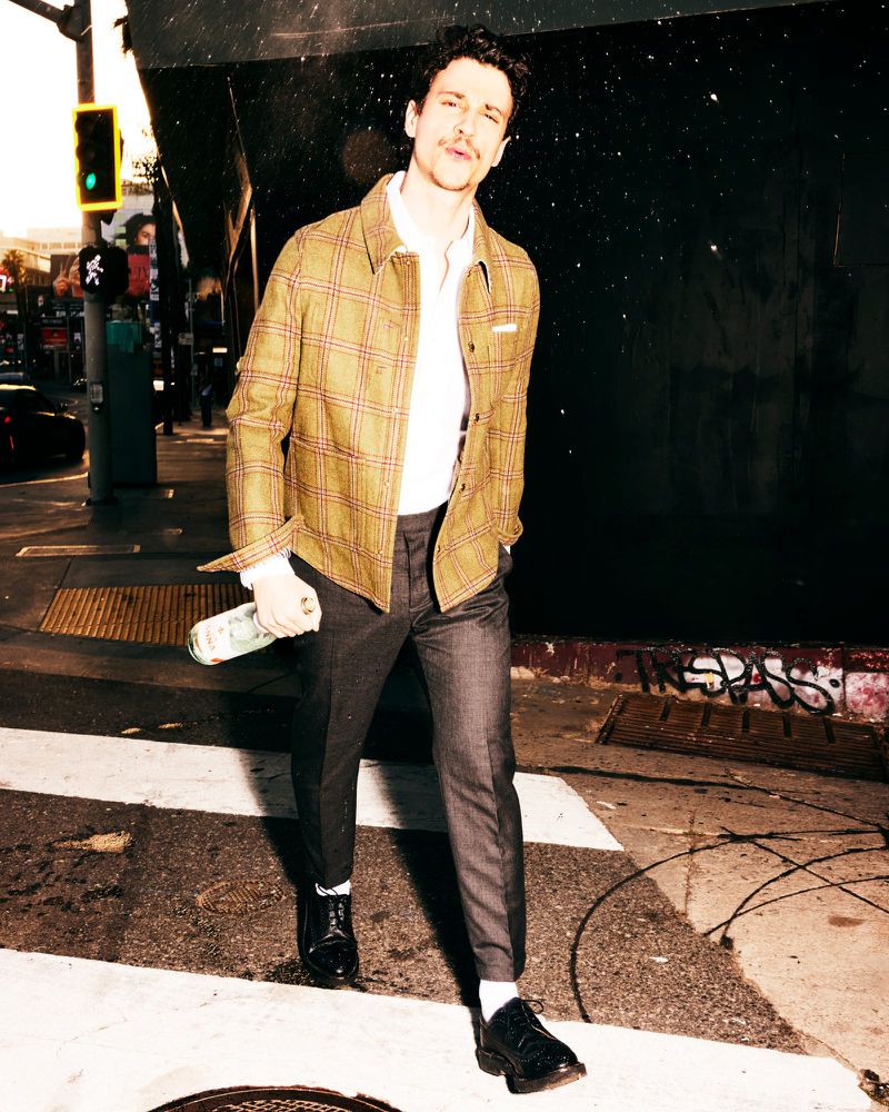 Man in jacket walks across crosswalk holding a bottle. He is wearing a white collared shirt, dark slacks, and dress shoes in outdoor shot.