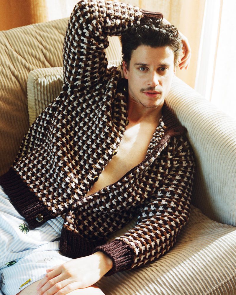 Adult male with facial hair lounges on a striped corduroy couch wearing a patterned sweater and pajama bottoms in a casual setting.