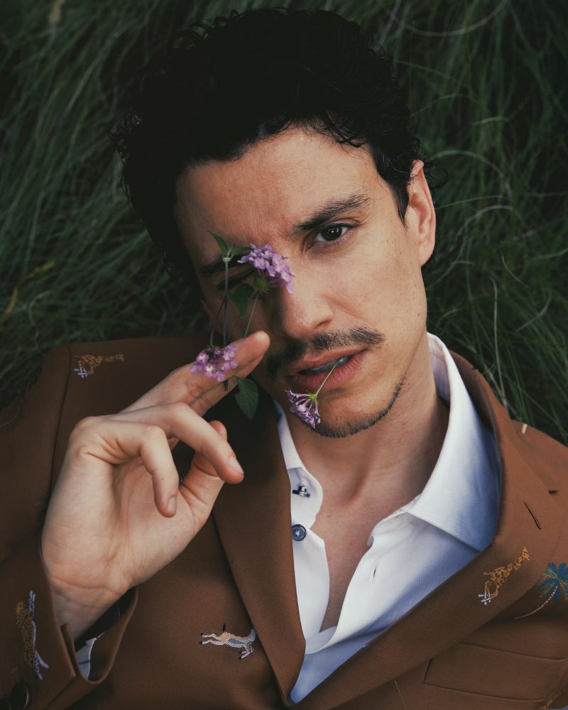 Portrait of a stylish adult man with dark hair and a mustache, laying in the grass with purple flowers, wearing a brown blazer with embroidery.