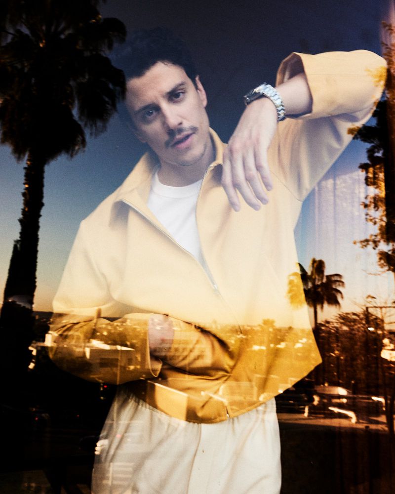 Fashion portrait of a young adult male with a mustache wearing a yellow jacket, with palm trees and an urban background in double exposure.