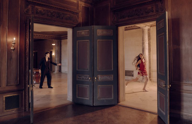 Elegant interior scene with a woman in a red dress running through a doorway, contrasted by a man in black standing in another.