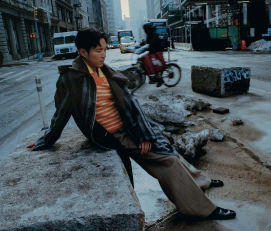Man in striped shirt and coat sitting on concrete block on urban street with city buildings and a cyclist in the background.