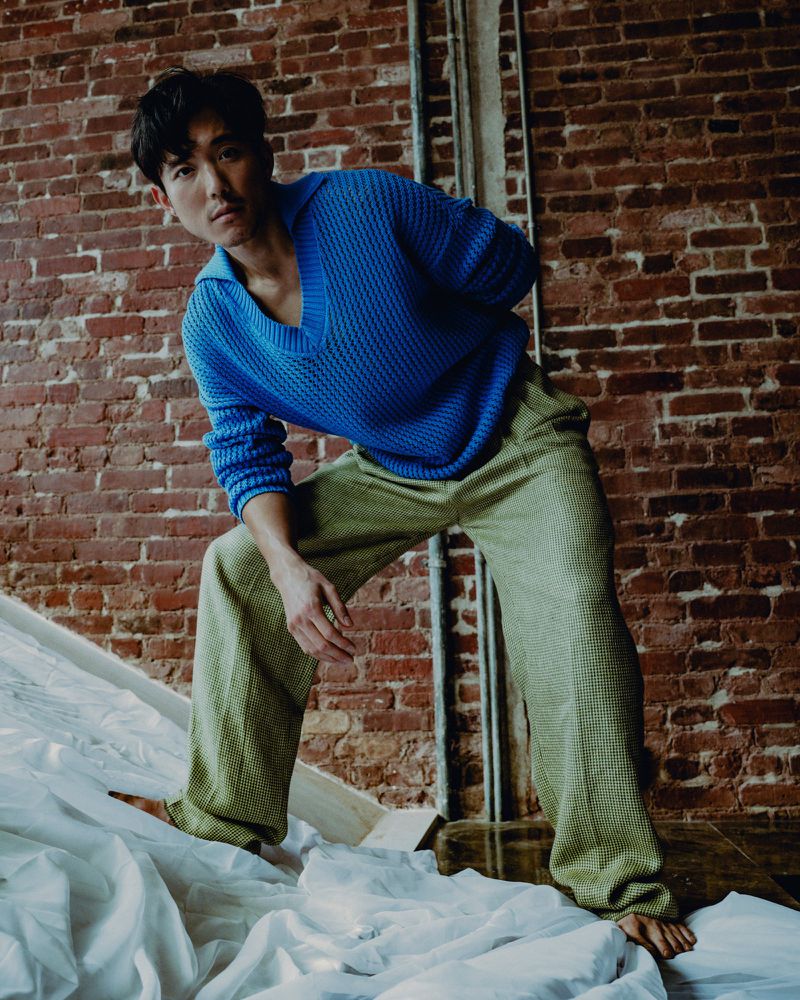 An Asian man poses in a vibrant blue sweater and patterned olive pants on a white cloth, set against a textured brick background indoors.