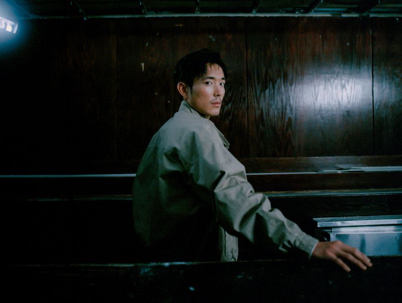 An adult male in a tan jacket is looking over his shoulder while sitting in a dark, wooden, church pew that is reflecting light.