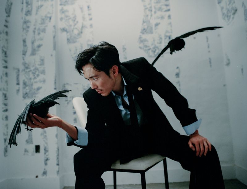 Asian man in suit sits holding crow, with another crow in the background, set against an artistic wall in a bright indoor setting.