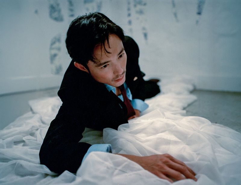 Asian man in a suit laying on a white cloth, captured in a studio setting. He is looking up with a subdued expression.