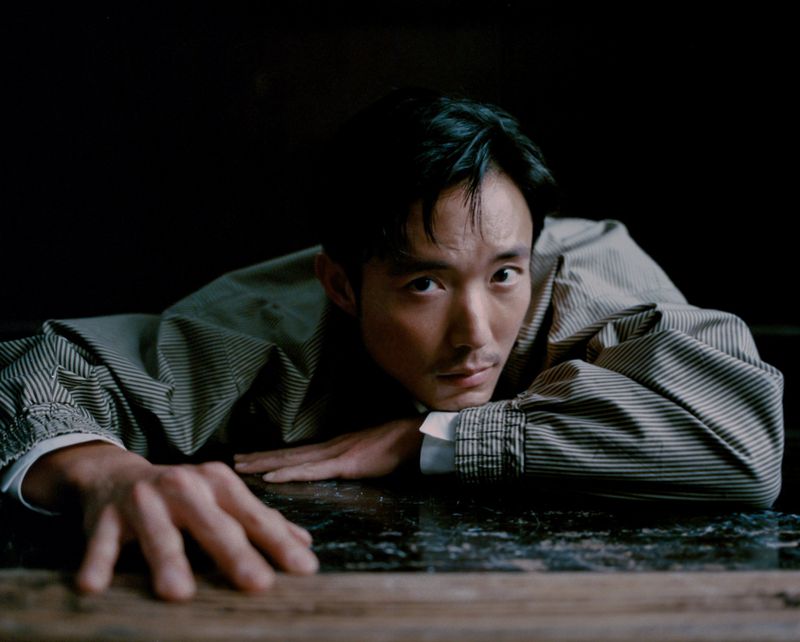 Portrait of a pensive Asian adult male in a striped jacket, laying with his hands flat on a reflective surface indoors against a dark backdrop.