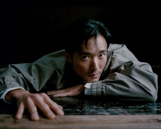 Portrait of a pensive Asian adult male in a striped jacket, laying with his hands flat on a reflective surface indoors against a dark backdrop.