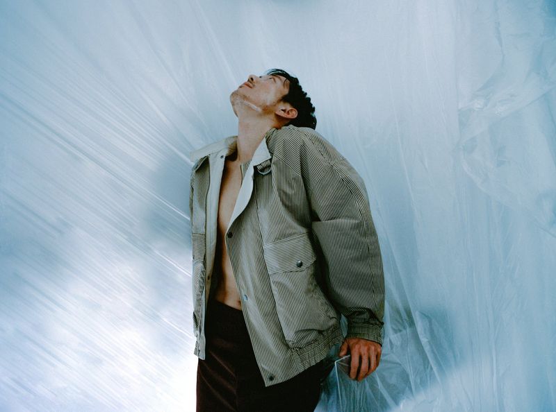 Stylish man in jacket looking up, surrounded by a translucent plastic sheet, captured in a fashion-forward, high-angle studio shot.