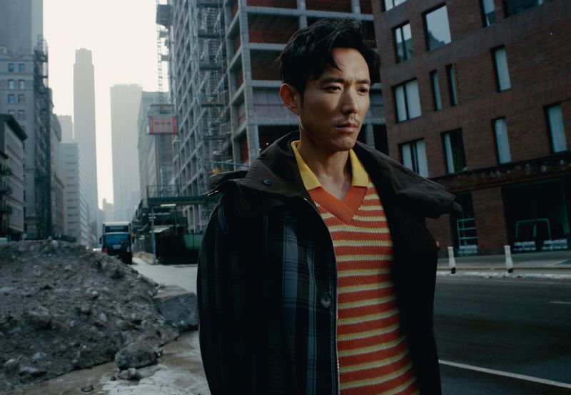 Man in a striped sweater and jacket stands on a city street in front of buildings, construction, and a blue bus.