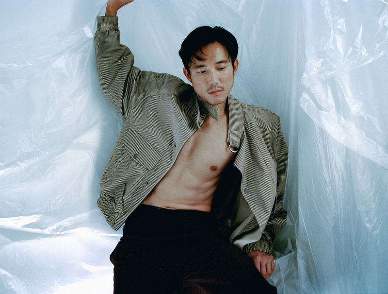 A shirtless Asian male model poses in a photo studio. He is wearing a striped jacket and black pants, with plastic sheeting in the background.