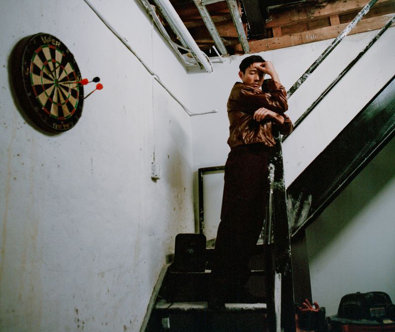 Man on stairs in brown jacket and pants. Dartboard visible on white-painted wall. Indoor shot with dim lighting.